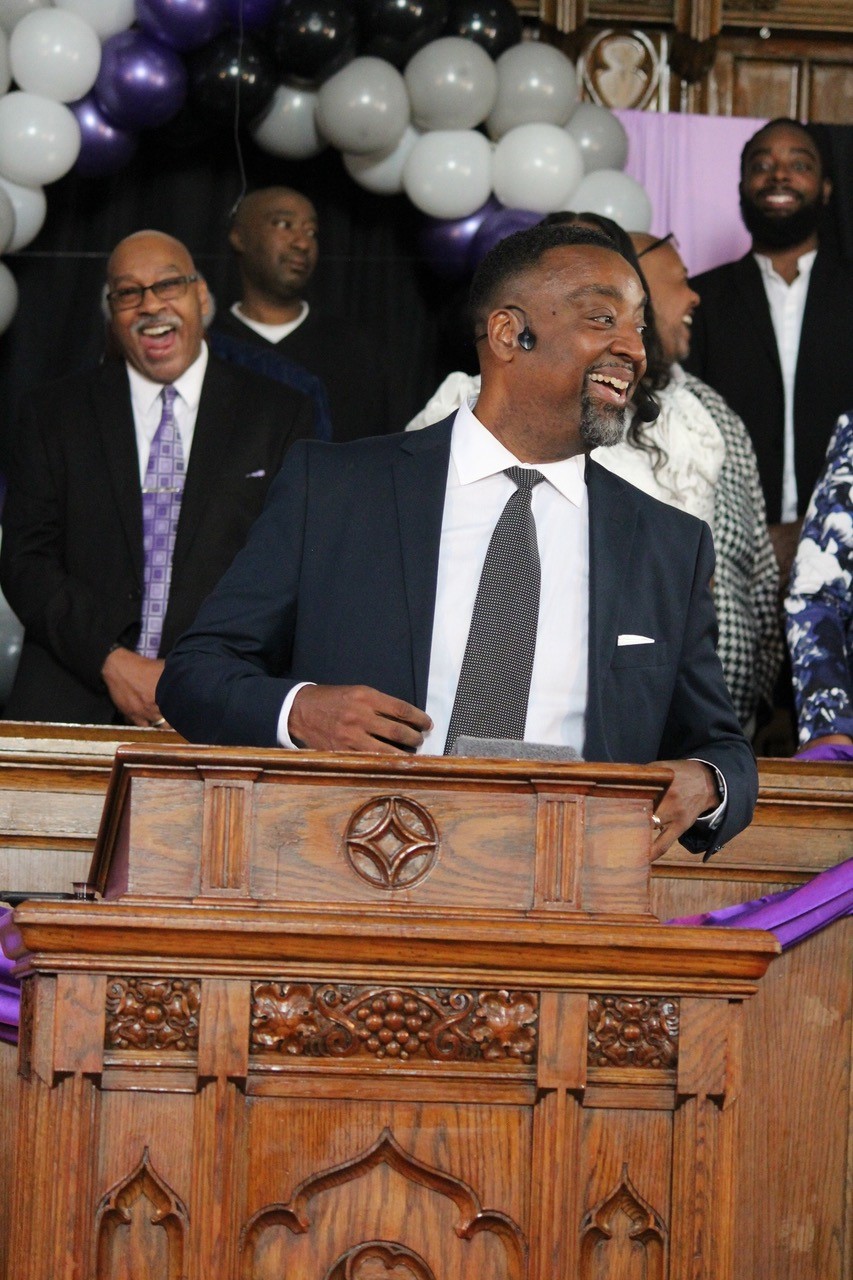 Pastor Steve Epting, in a black suit, white shirt and gray tie, speaks to the congregation with a smile on his face while standing at a wooden pulpit.