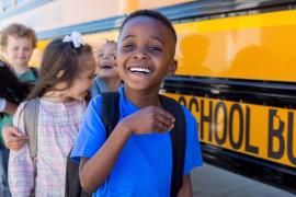 A young boy with a backpack smiles at the front of a line of children waiting to board a yellow school bus.