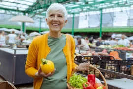 A senior woman holds a pepper and basket of groceries at a busy supermarket.