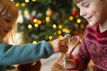 Two kids sitting under a Christmas tree hold up candy canes in the shape of a heart.