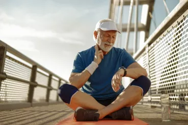 A senior man in jogging gear sits cross-legged on a bridge and checks his pulse while looking at his watch.