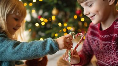 Two kids sitting under a Christmas tree hold up candy canes in the shape of a heart.