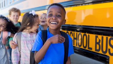 A young boy with a backpack smiles at the front of a line of children waiting to board a yellow school bus.