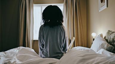 A woman sitting up in bed facing a window.