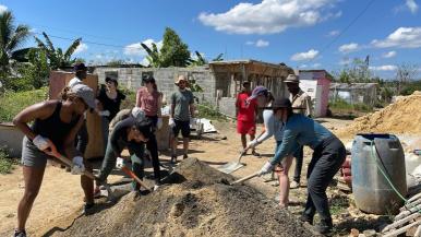 Students from the RISE program shovel dirt in front of a home in the Dominican Republic on a sunny day.