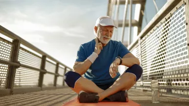 A senior man in jogging gear sits cross-legged on a bridge and checks his pulse while looking at his watch.