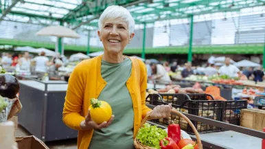 A senior woman holds a pepper and basket of groceries at a busy supermarket.