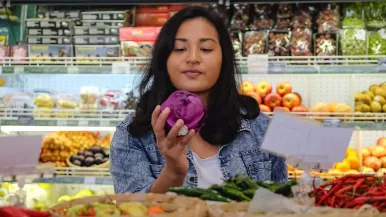 A young woman considers a vegetable she has picked out in the produce aisle of a grocery store.