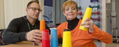 A male physical/occupational therapist works with a woman patient on fine motor skills. They sit at a table with colorful cones, and the patient holds a yellow cone up.